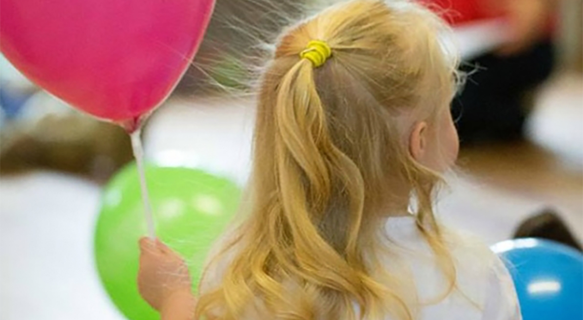 Girl with her back to the camera with blonde hair in ringlets, holding a pink balloon.