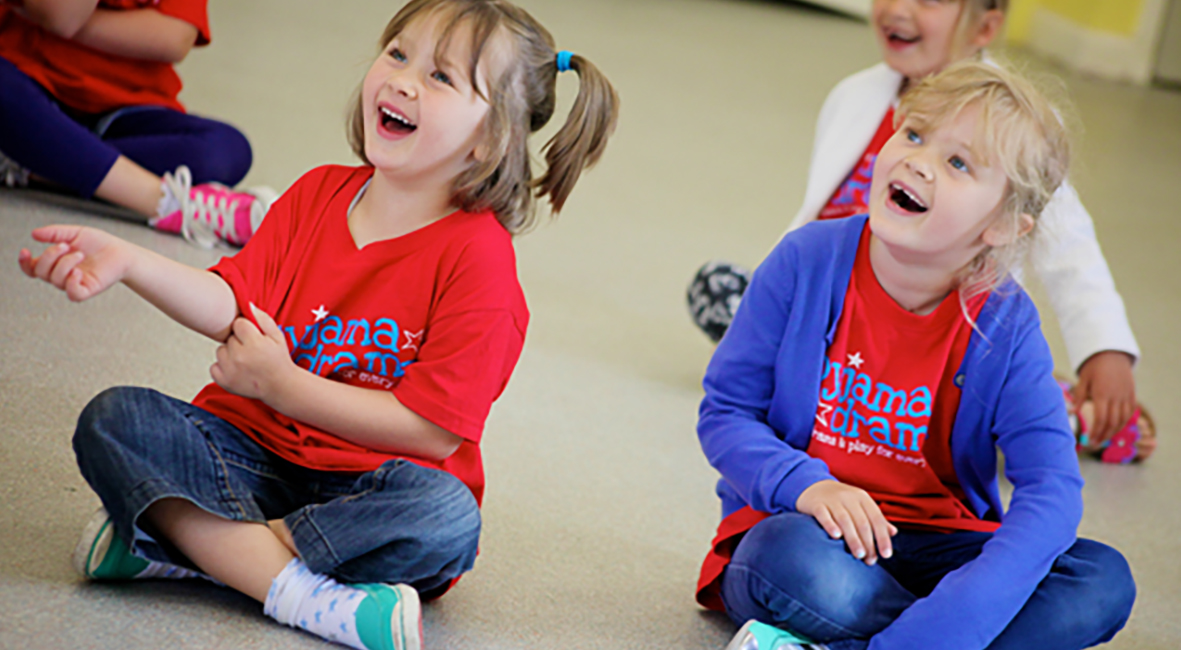 Two girls sit on the floor in a Pyjama Drama class laughing at something they can see off shot.